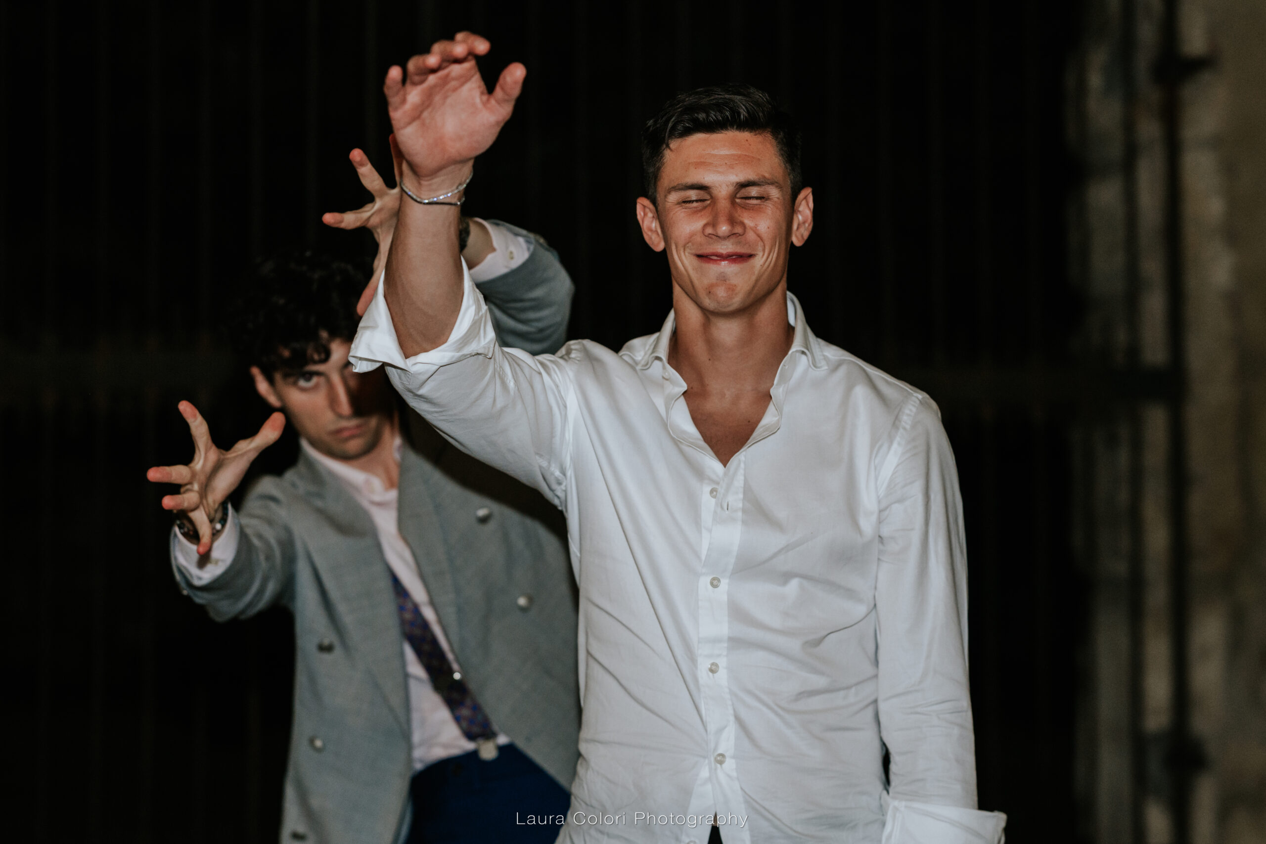 A man in a white shirt smiles with eyes closed as he raises his hand; another man in a gray blazer gestures playfully behind him at night.