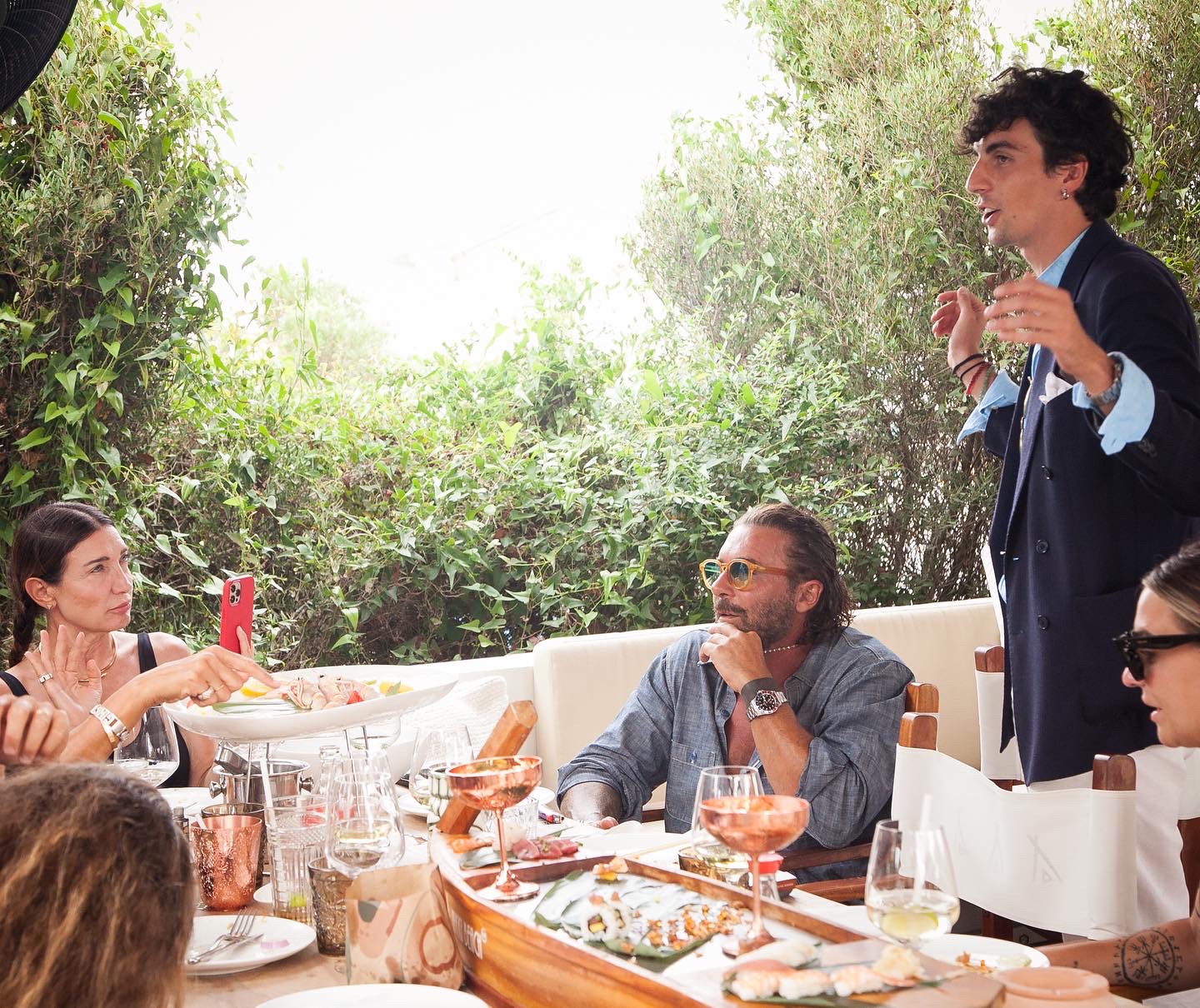 Group of friends at an outdoor lunch, a man in a dark blazer stands and gestures as others listen at the long table filled with sushi and drinks.