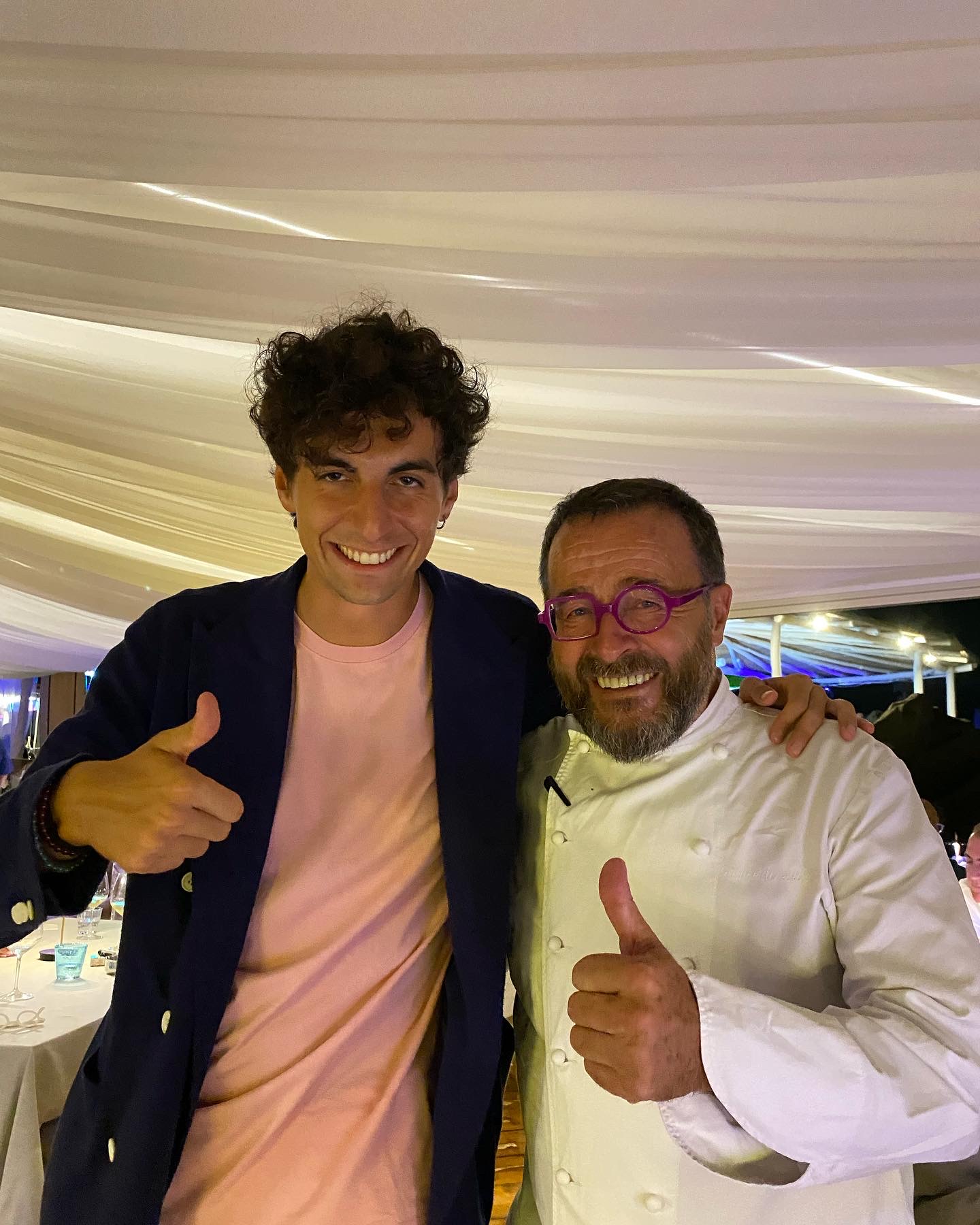 Two men pose together at a celebration, both giving a thumbs up and smiling; one in a pink shirt with a navy blazer, the other in a white chef's coat with purple glasses.